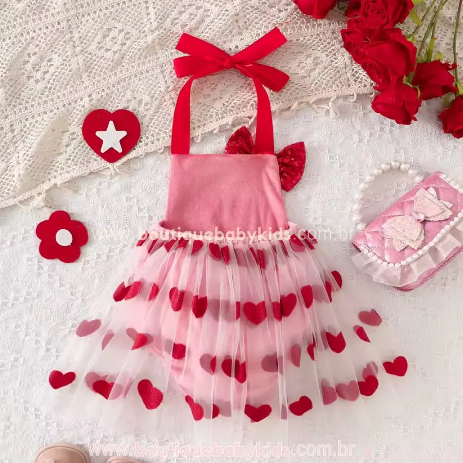 Vista traseira do body bebê rosa com saia de tule de corações vermelhos, exibido em um cenário decorado com acessórios delicados, ideal para ensaios fotográficos e datas comemorativas.