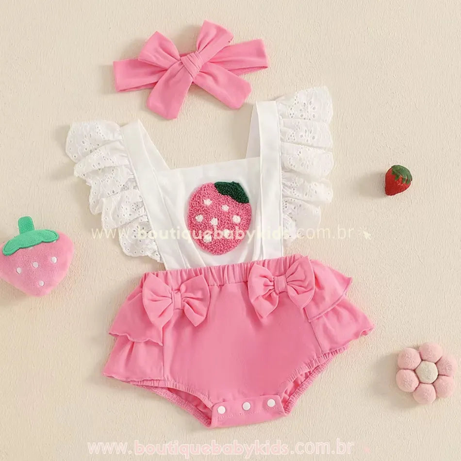 Conjunto do romper bebê rosa bordado com morango ao lado de acessórios temáticos, incluindo uma faixa de cabelo rosa, um enfeite de morango de pelúcia e detalhes decorativos suaves.