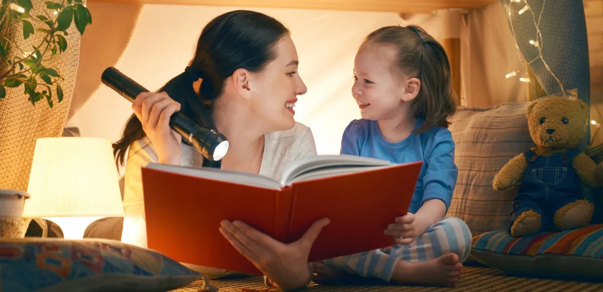 Mãe e filha em um ambiente aconchegante lendo um livro juntas, iluminadas por uma lanterna, criando um momento especial de leitura noturna.
