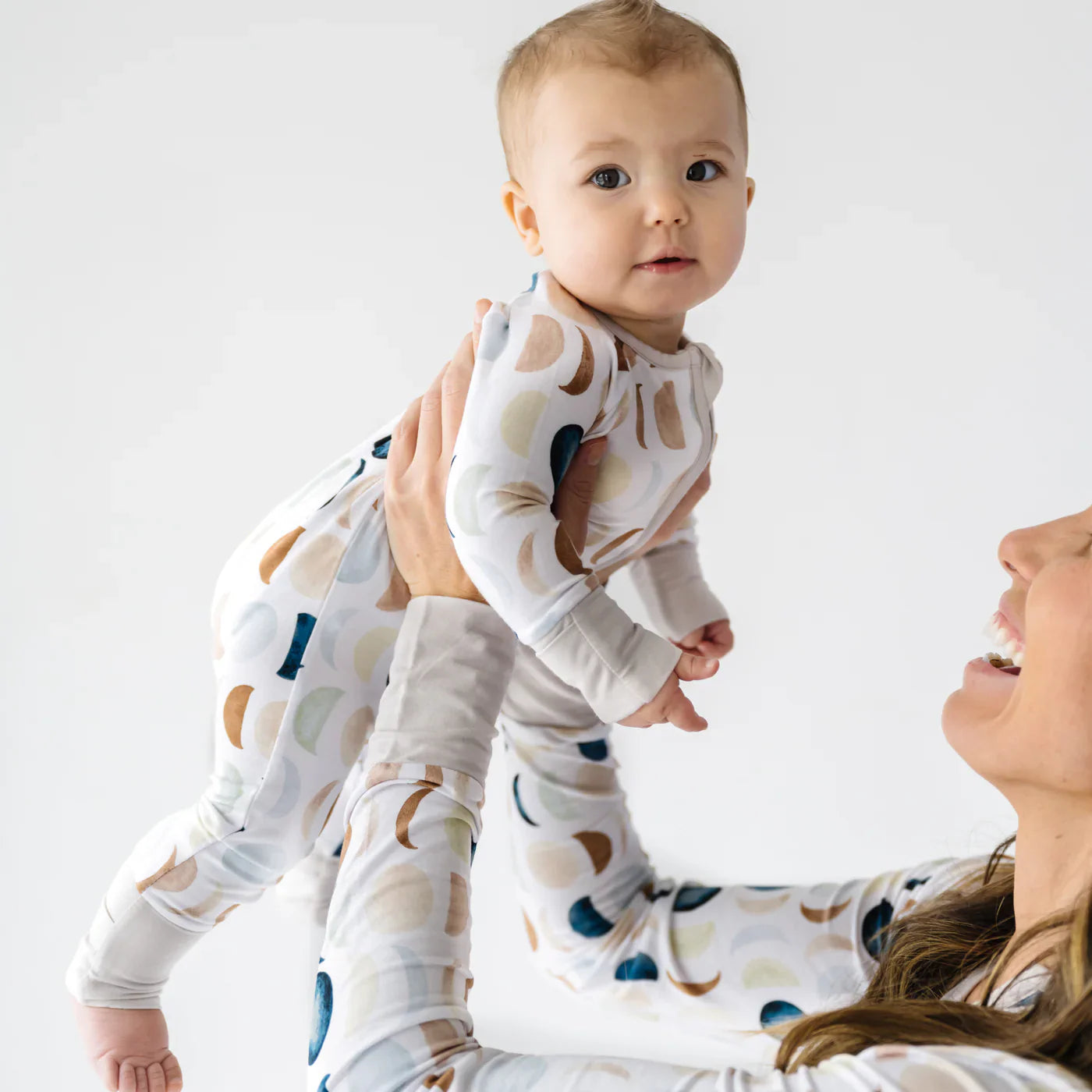 Bebê usando macacão de bambu com estampa de luas em tons neutros, sendo levantado pela mãe com roupa combinando.