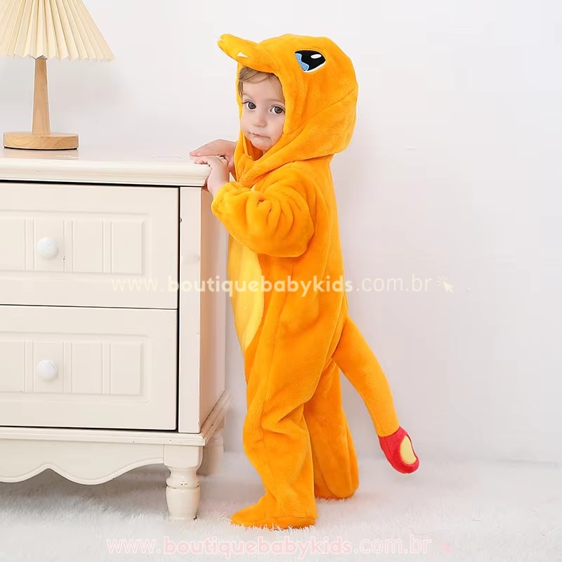 Bebê em pé ao lado de um móvel vestindo o Macacão Bebê Fantasia Pokémon Charmander, destacando o tecido peluciado e a cauda longa.