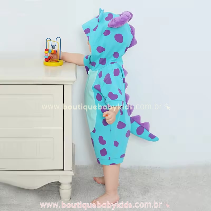 Bebê vestindo o Macaquinho Bebê Fantasia Sulley Monstros S.A, explorando um móvel branco, destacando o design fofo com capuz de chifrinhos e cauda temática.