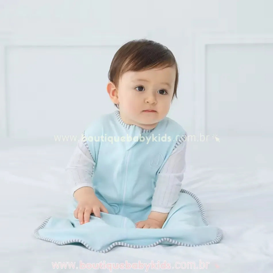 Bebê sentado sobre a cama, usando o saco de dormir azul claro com bordas listradas e zíper frontal, com um olhar curioso.