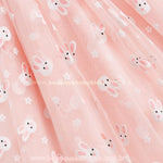 Close na saia do vestido rosa, evidenciando o tule leve e fluído com estampa de coelhinhos brancos e detalhes de pequenas flores, ideal para um visual delicado e festivo. 