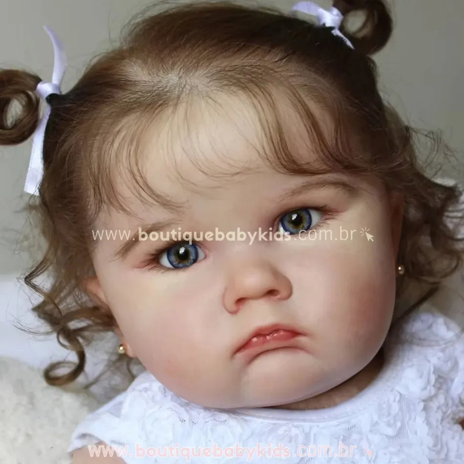 Close do rosto da Boneca Bebê Reborn Camila com laços brancos no cabelo castanho encaracolado, mostrando os olhos castanhos e expressão delicada.