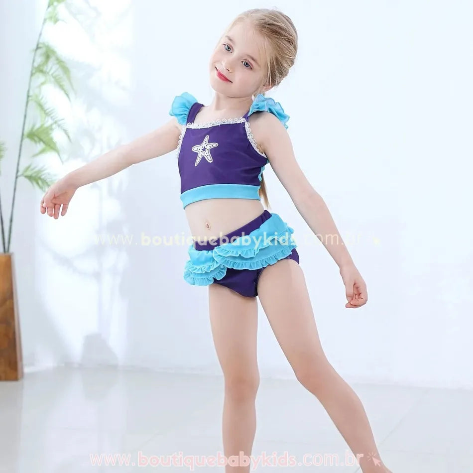 Menina sorridente vestindo o Biquíni Infantil Princesa Ariel, em pose graciosa, destacando o top lilás com aplicação de estrela-do-mar e babados azul-claros nos ombros, combinando com a calcinha lilás adornada com camadas de babados delicados.