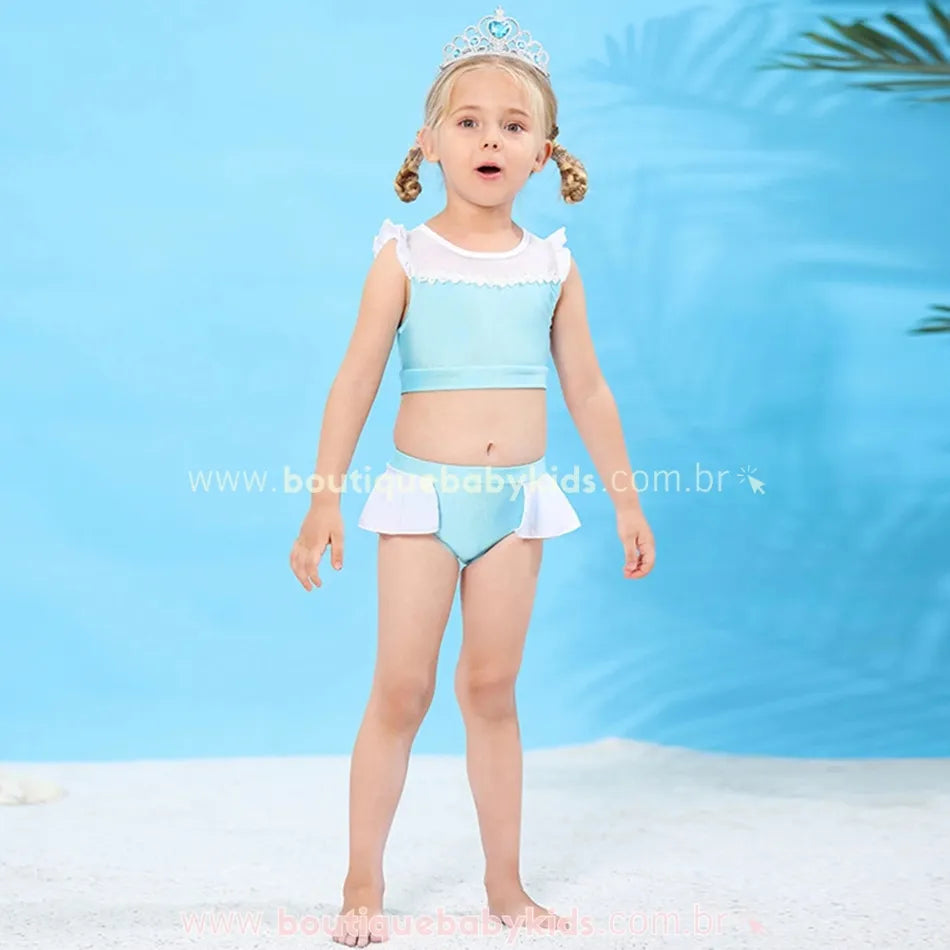 Menina com biquíni temático da Cinderela, em pé, expressando surpresa, destacando os detalhes encantadores do conjunto em um ambiente de praia artificial.