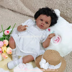 Boneca Reborn Cecília de pele negra deitada sobre tapete felpudo bege, vestindo vestido branco de renda, segurando borboleta decorativa.