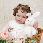 Close-up do rosto da Boneca Bebê Reborn mostrando detalhes dos olhos verdes e cabelos castanhos cacheados, segurando um coelho de pelúcia branco próximo à bochecha.