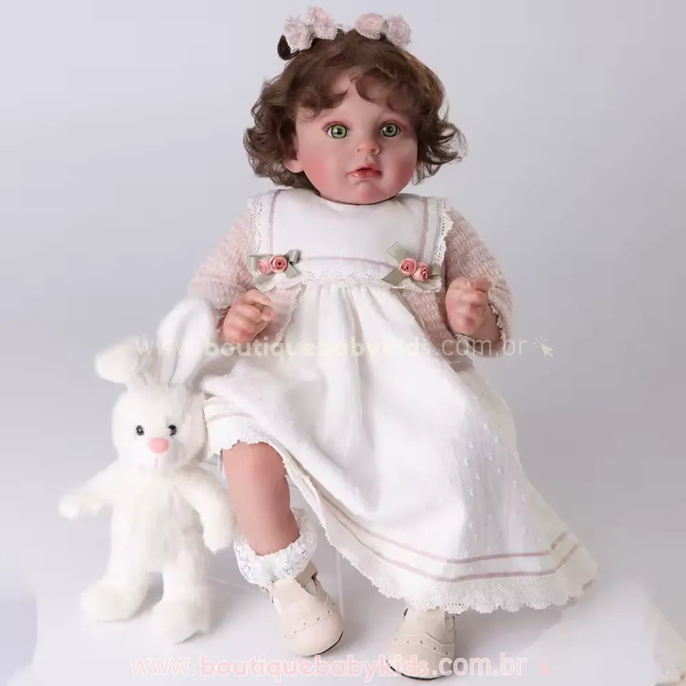 Boneca Bebê Reborn realista sentada em fundo branco, vestindo vestido branco com casaco de tricô rosa e meias de renda, acompanhada de um coelho de pelúcia branco ao lado.