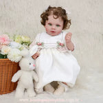 Boneca realista estilo Reborn sentada ao lado de uma cesta de vime com flores artificiais coloridas, vestindo conjunto clássico branco e rosa, com coelho de pelúcia.