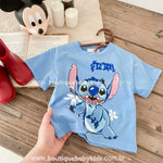 Camiseta azul infantil com estampa do Stitch, sendo segurada por uma mão, com fundo de madeira, livro aberto, bota vermelha e enfeite do Mickey Mouse ao fundo.