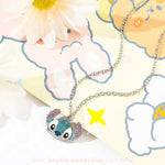 Colar infantil com pingente do Stitch cravejado com strass azul e rosa, sobre fundo decorado com desenhos coloridos e flor branca.