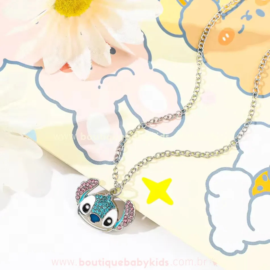 Colar infantil com pingente do Stitch cravejado com strass azul e rosa, sobre fundo decorado com desenhos coloridos e flor branca.