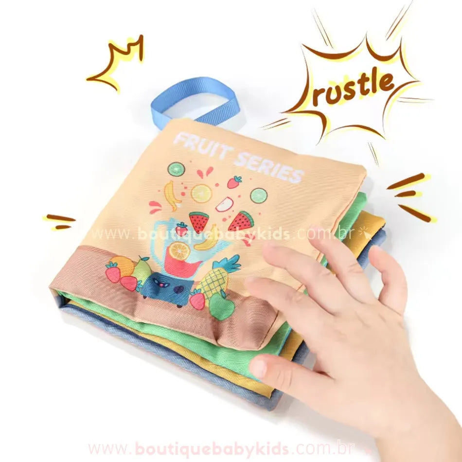 Mão infantil tocando um dos livros sensoriais, ativando um efeito sonoro ao virar a página. Ilustração mostra frutas como laranja e melancia.