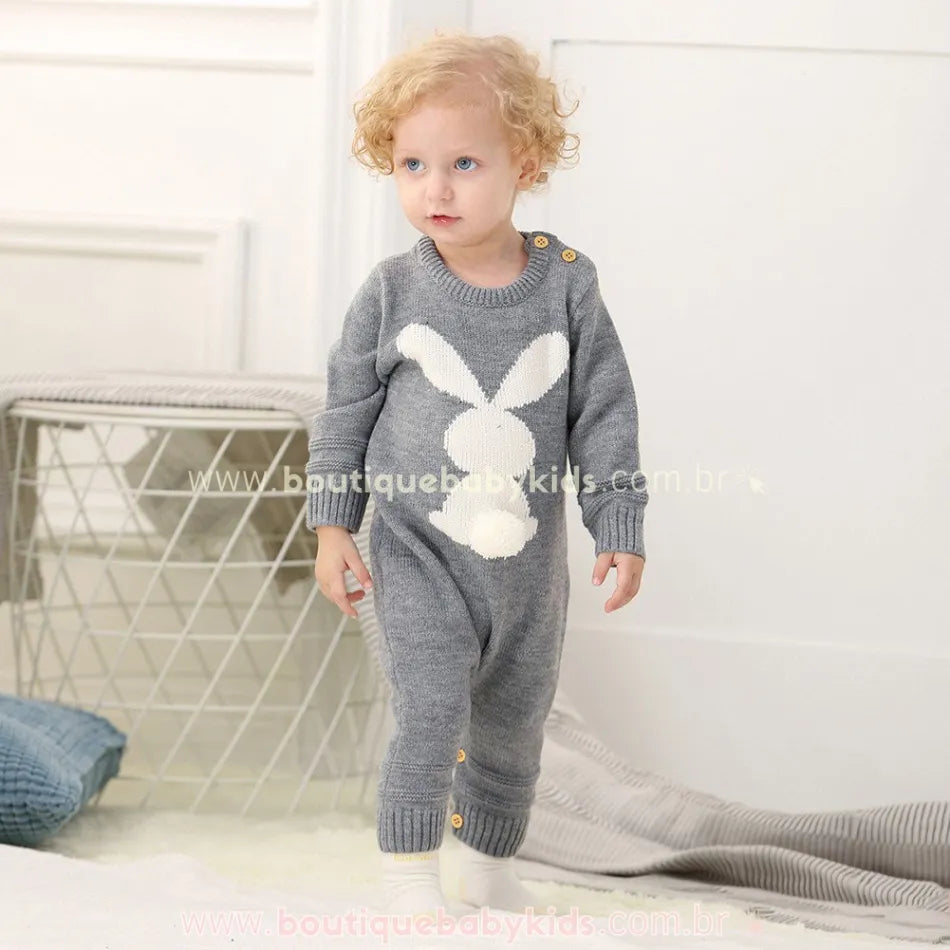 Bebê andando pelo ambiente aconchegante, vestindo o Macacão Bebê Tricot Coelhinho cinza. O macacão conta com fechamento por botões na lateral do ombro e entre as pernas. 