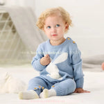 Bebê de olhos claros sentado no chão, vestindo o Macacão Bebê Tricot Coelhinho azul. O macacão tem um coelhinho branco bordado e botões de madeira decorativos. 