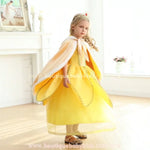 Menina vestida como uma princesa usa a capa infantil dourada com acabamento em pelúcia branca. Ela gira suavemente em um ambiente elegante, destacando o caimento fluído da peça e os detalhes sofisticados do tecido aveludado. O capuz e o laço frontal complementam o visual encantador, tornando a capa perfeita para ocasiões especiais e fantasias temáticas. 